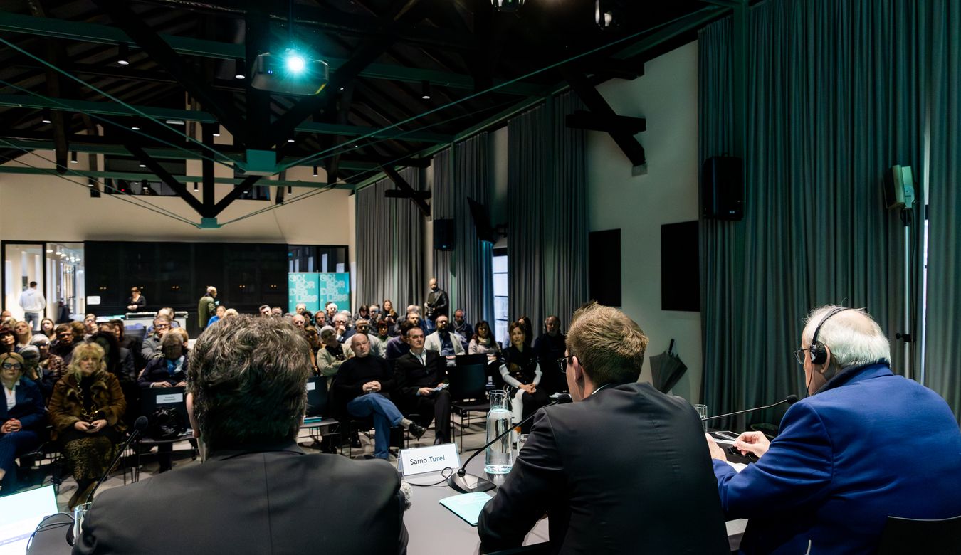 Fotografija z govorniškega pulta proti sedeči publiki v dvorani.